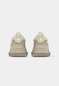 Beige athletic shoes with a textured mesh design, rounded shape, soft fabric accents, and pull tabs on the back. Subtle tonal detailing throughout.