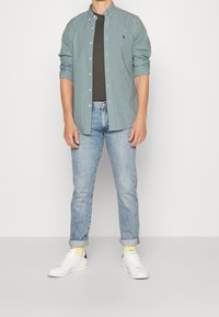 Green and white checkered shirt, unbuttoned over a dark green t-shirt, paired with light blue jeans and white sneakers with yellow accents.