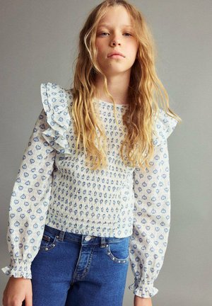 Blond meisje met lang golvend haar, gekleed in een witte blouse met blauwe bloemenpatronen en gerimpelde mouwen, gecombineerd met blauwe jeans.