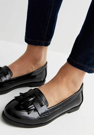 Sorte patentlæder loafers med frynser, båret med mørkeblå jeans på bare fødder foran en lys baggrund.