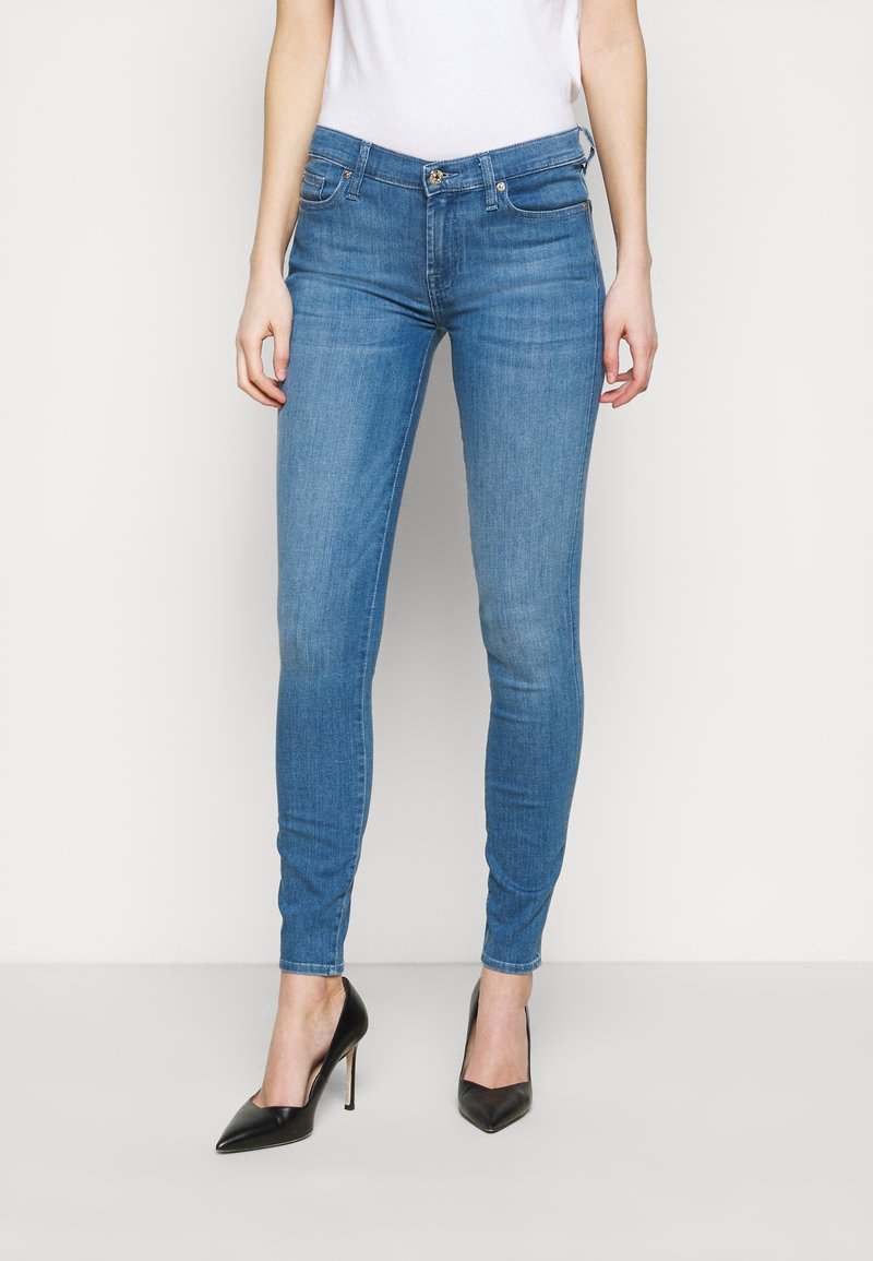 Vaqueros ajustados de denim azul, con un diseño clásico de cinco bolsillos, cierre de cremallera y botón, combinados con zapatos de tacón negros.