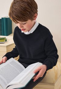Pull noir en maille avec un col rond, porté par-dessus une chemise blanche à col. La personne tient un livre ouvert sur une surface en bois clair.