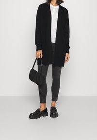 Lång svart cardigan i mjukt stickat material, tillsammans med en vit skjorta och mörkgrå skinny jeans. Tillbehören inkluderar en texturerad svart väska och kraftiga skor.