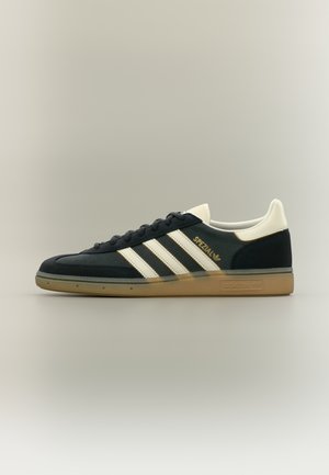 Zapatilla negra de ante con detalles en cuero blanco, con tres franjas blancas y la marca "Spezial" en dorado. Suela de goma marrón.