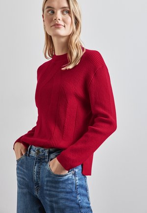 Maglione rosso a coste con collo rotondo, caratterizzato da motivi verticali e spacchi laterali. Abbinato a jeans in denim blu.