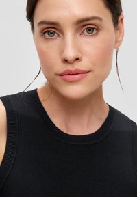 Zwarte mouwloze gebreide top met een ronde halslijn en geribbelde randen, gedragen door een model met subtiele make-up en steil haar tegen een grijze achtergrond.