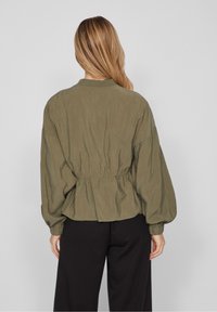 Oliwkowa krótka kurtka z elastycznym paskiem w talii, bufiastymi rękawami i kołnierzykiem. Lekki, pomarszczony materiał i proste detale w projektowaniu.