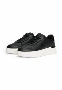 Un paio di sneakers in pelle nera con suole bianche spesse, lacci neri e un logo in rilievo discreto sul lato, fotografati su uno sfondo bianco.