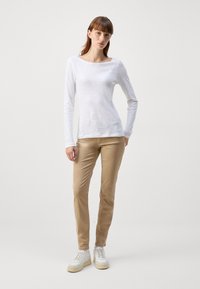 Långärmad vit topp med båthalsringning, matchad med beiga rakbenade byxor. Modellen bär vita sneakers med beige detaljer.