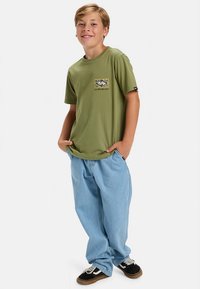 Žalia trumpomis rankovėmis marškinėliai su grafiniu kišenės dizainu, derinami su šviesiai mėlynais laisvesniais džinsais ir juodais sportbačiais su rudos spalvos pado detalėmis.