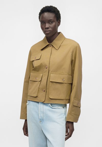 Γυναίκα που φοράει ένα μπεζ μπουφάν με κουμπιά και τέσσερις τσέπες με καπάκια και ανοιχτό μπλε τζιν, στέκεται μπροστά σε απλό λευκό φόντο.