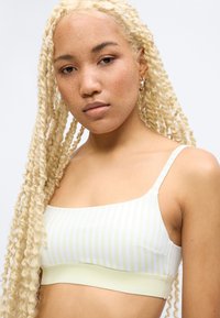 Νέα γυναίκα με μακριές ξανθές πλεξίδες, που φοράει ένα κοντό τοπ με λεπτές τιράντες σε απαλό κίτρινο και άσπρες ρίγες, κοιτάζει ήρεμα την κάμερα.