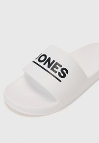 Sandale blanche avec une large bride portant une impression noire "JONES". Texture lisse et mate avec un support pour le pied en contours et une semelle plate.