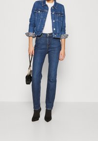 Chaqueta de denim con puños enrollados sobre una camisa blanca, combinada con jeans azul oscuro y botas negras de punta. Bolso negro con cadena.