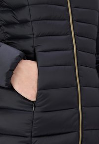 Veste matelassée noire avec texture matelassée, fermeture éclair dorée et poches latérales. Le matériau lisse avec un design ajusté met en valeur la chaleur et la fonctionnalité.
