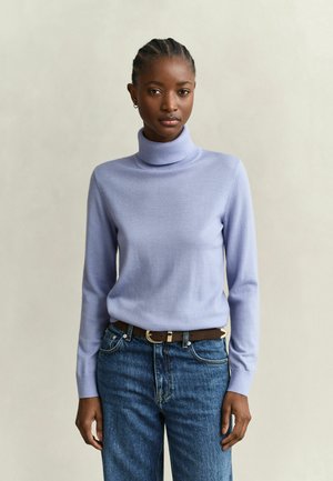 Femme aux cheveux tressés portant un pull col roulé violet clair et un jean bleu avec une ceinture marron, debout devant un fond clair uni.