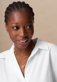 Zlati uhani, vključno s pendantom in verigo, so položeni čez belo srajco s ovratnikom. Subjekt ima pletene lase in naravno ten kože.