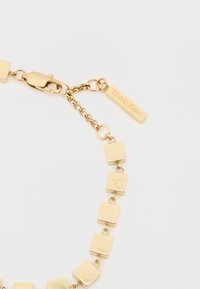 Χρυσό βραχιόλι με τετράγωνα συνδέσμους, κλείσιμο με κούμπωμα και ετικέτα Calvin Klein. Λείος επιφανειακός με γυαλιστερό φινίρισμα.