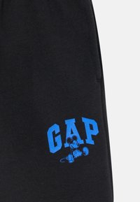 Pantalons de survêtement noirs avec un logo "GAP" bleu et une illustration d'une souris de dessin animé, fabriqués à partir d'un mélange de coton doux et texturé.