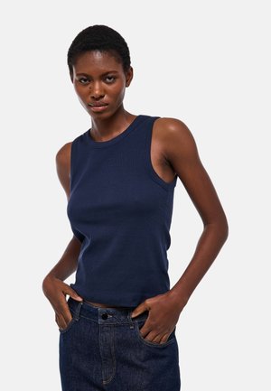 Femme portant un haut sans manches bleu marine et un jean foncé, debout les mains dans les poches devant un fond uni clair.