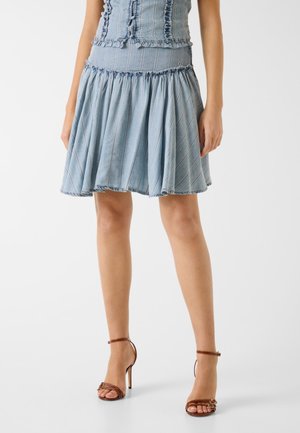 Kobieta ubrana w jasnoniebieską, rozkloszowaną do kolan spódnicę jeansową z pionowymi przeszyciami oraz brązowe sandały na wysokim obcasie z paskami wokół kostki.