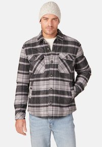 Grijze en zwarte geruite flanellen jas met paarse accenten, knopen aan de voorkant, twee borstzakken, gedragen over een crèmekleurige trui.