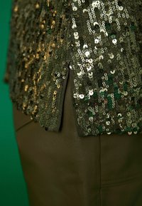 Vêtement vert à paillettes avec une surface texturée, présentant des paillettes qui se chevauchent dans différentes nuances de vert et d'or, associé à un matériau lisse.