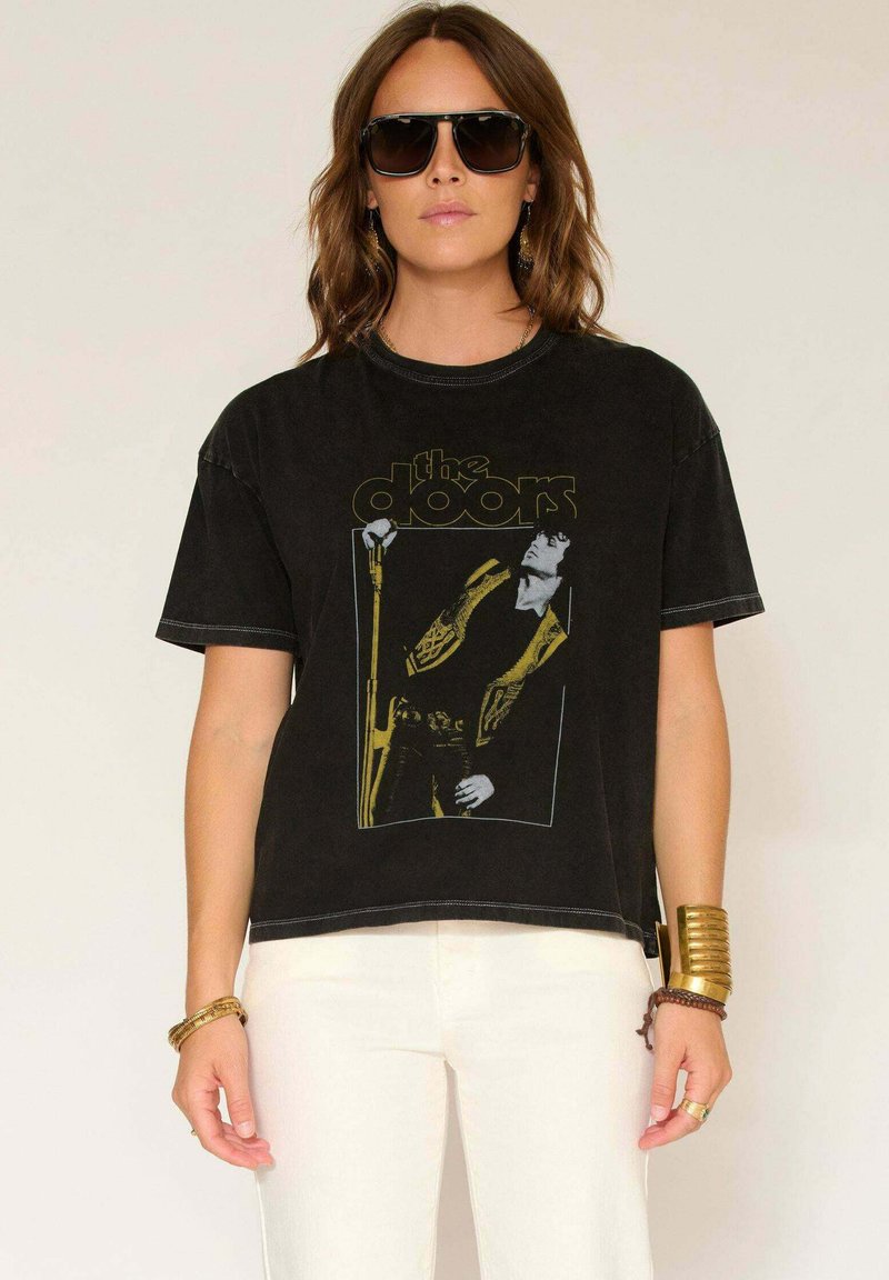 Femme portant un t-shirt noir du groupe The Doors, un pantalon blanc, de grandes lunettes de soleil noires et des bracelets en or, se tenant devant un fond uni.