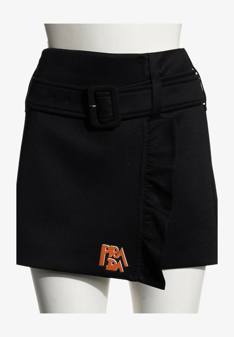 Jupe courte noire avec ceinture, détail à volants sur le devant, et logo Prada orange près de l'ourlet du côté gauche.