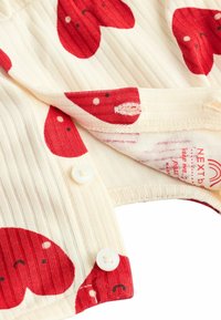 Crème ribstof met rode hartpatronen, voorzien van witte knopen en een label met verzorgingsinstructies en merknaam.