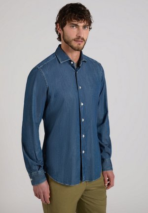 Mężczyzna z kręconymi włosami, ubrany w zapinaną na guziki niebieską koszulę z denimu i oliwkowe spodnie, stoi na tle jednolitego jasnego tła.