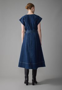 Vestido de mezclilla en azul oscuro con mangas cortas, cintura ajustada y falda acampanada. Detalles de costura visibles y textura suave. Botas negras hasta la rodilla.