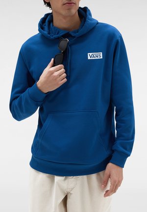 Persona che indossa una felpa blu Vans, tiene occhiali da sole neri, abbinata a pantaloni beige chiaro, su uno sfondo bianco semplice.