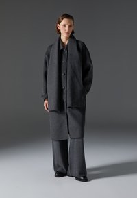 Abrigo de lana gris con botones grandes, combinado con pantalones amplios grises. La textura suave y el corte holgado realzan el diseño minimalista.