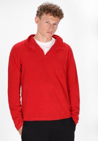 Rød fleece pullover med en kvart lynlås krave, lange ærmer, blød tekstur og en afslappet pasform. Båret over en hvid skjorte, med sorte bukser.