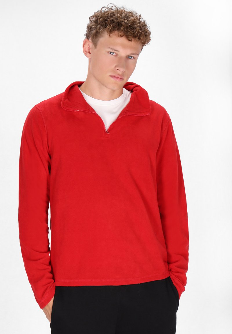 Rød fleece pullover med en kvart lynlås krave, lange ærmer, blød tekstur og en afslappet pasform. Båret over en hvid skjorte, med sorte bukser.