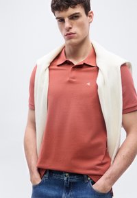 Νεαρός άνδρας φοράει πορτοκαλοκαφέ πόλο μπλουζάκι με ένα λευκό πλεκτό πουλόβερ περασμένο στους ώμους του, χέρια στις τσέπες του μπλε τζιν.