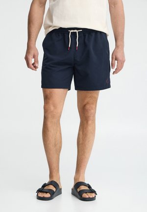 Hombre con pantalones cortos azul marino con cordón y sandalias negras con hebilla, de pie contra un fondo claro y liso.