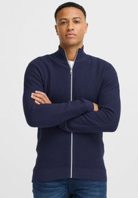 Námornícky zipsový sveter s textúrovaným pletením, vysokým golierom a kontrastným strieborným zipsom. Nosený cez biele tričko a spárovaný s modrými džínsami.