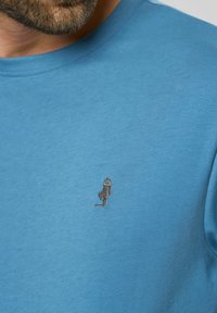 Kék pamut póló rövid ujjakkal, a bal mellrészen egy kis, részletes fekete hímzett figura található. Simaság jellemzi.