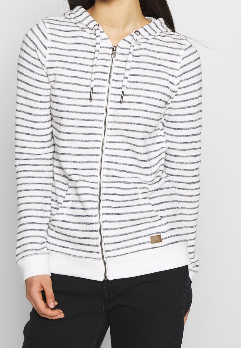 Femme portant un sweat à capuche blanc zippé à fines rayures horizontales foncées et un pantalon noir, debout devant un fond clair uni.