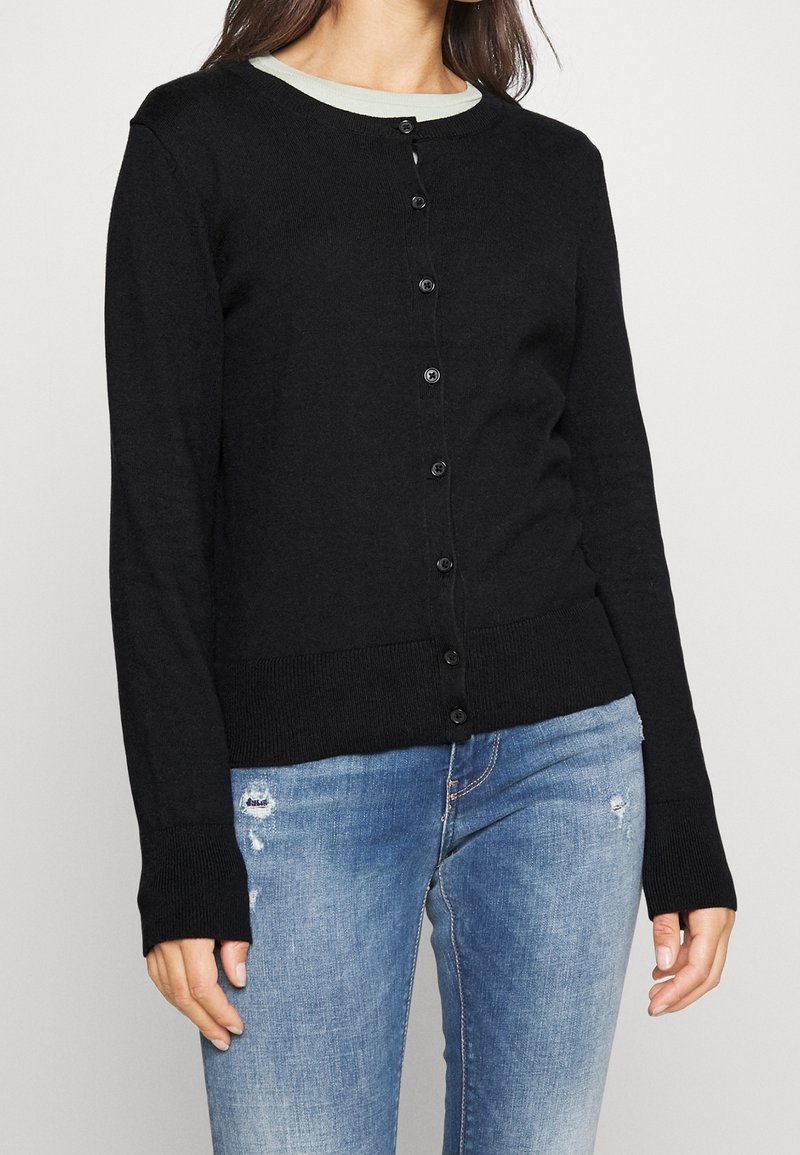 Femme portant un cardigan noir boutonné sur une chemise claire et un jean bleu clair délavé, debout, les mains détendues le long du corps.