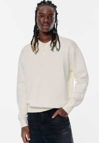 Cremfarbener Pullover mit Knopfkragen, gerippten Bündchen und weicher Textur, kombiniert mit dunklen Jeans.