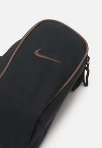 Černý látkový sáček s hladkým povrchem, zdobený bronzovým logem Nike a uzávěrem na zip. Tvarovaná silueta s tmavými akcenty.