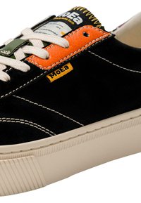 Juodas drobės sportbačių modelis su baltais siūlais, tekstūruotais oranžiniais ir žaliais akcentais, kreminiu guminiu padu ir pažymėtu liežuvėliu.