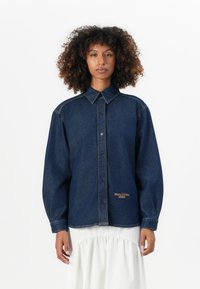 Vrouw met krullend haar draagt een donkerblauwe denim overhemd met knopen met het "Marc O'Polo Denim" logo en een witte, geplooide rok tegen een witte achtergrond.