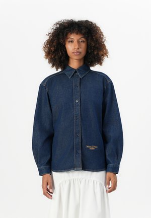 Vrouw met krullend haar draagt een donkerblauwe denim overhemd met knopen met het "Marc O'Polo Denim" logo en een witte, geplooide rok tegen een witte achtergrond.