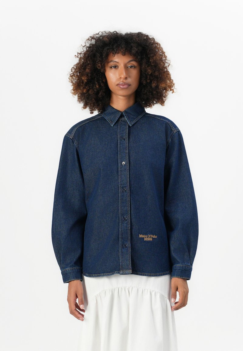 Vrouw met krullend haar draagt een donkerblauwe denim overhemd met knopen met het "Marc O'Polo Denim" logo en een witte, geplooide rok tegen een witte achtergrond.