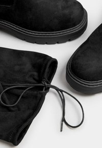 Svarta mocka ankelstövlar med texturerad yta, tjocka svarta gummisulor och dragsnodddetalj från en matchande svart mockaväska.