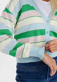 Kvinna som bär en randig kofta i pastellblått, grönt, rosa och vitt med knappar, som justerar ärmslutet, tillsammans med blå denimjeans.